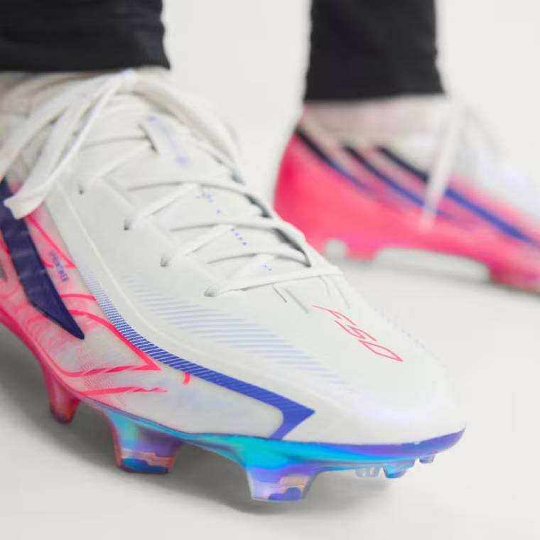 Близък план на бели футболни обувки adidas F50 Hyperfast Elite FG с розови графични детайли, сини акценти и цветна блестяща подметка с бутони за терен със суха естествена трева 