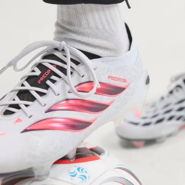 Близки план на бели футболни бутонки adidas Predator с ярки розово-червени ивици, стъпили върху футболна топка